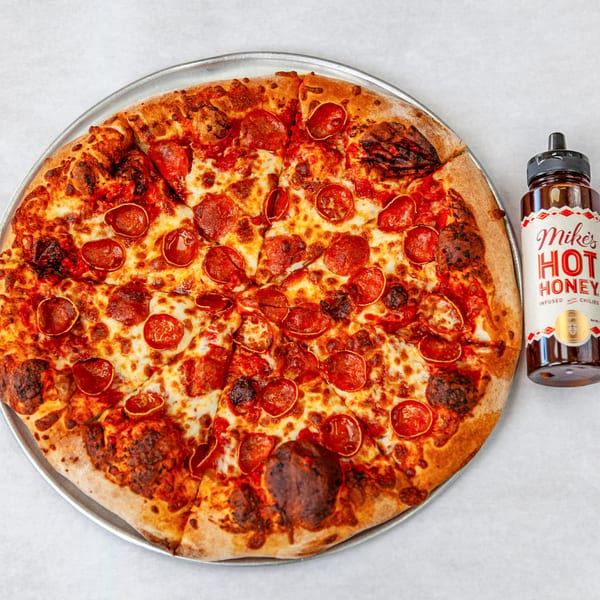 A large pepperoni pizza on a metal tray next to a bottle of Mike's Hot Honey.