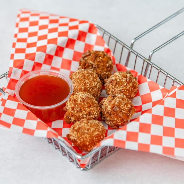 Six golden-brown fried appetizers and a side of red dipping sauce in a metal basket with checkered paper.