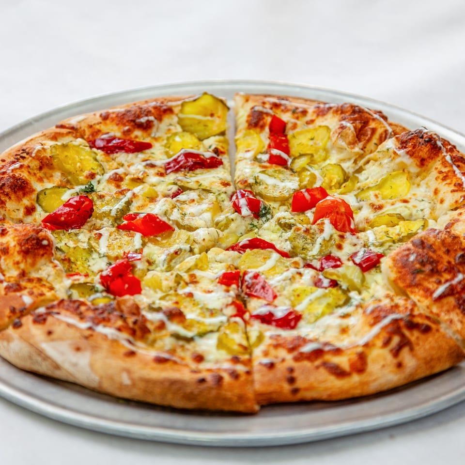 Home Slice Pizzeria's pizza with pickles, red peppers, and white sauce on a metal tray.